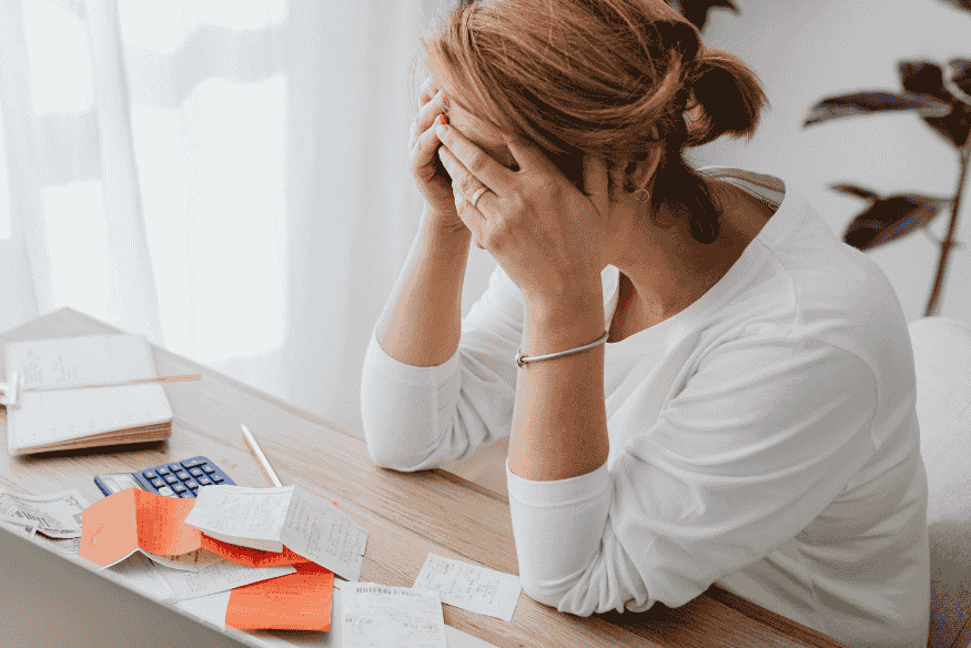 Donna seduta al tavolo con la testa china e la mano sulla fronte, circondata da documenti e bollette